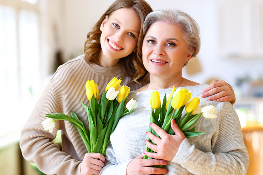 Positive young daughter with senior mother in casual clothes smiling and hugging while standing at home with bunches of fresh delicate tulips during celebration of Mother Day 